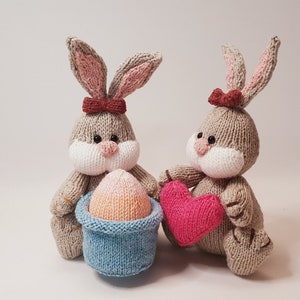 Peut inclure: Deux lapins en tricot, l'un tenant un cœur rose et l'autre tenant un panier bleu avec un œuf rose et orange à l'intérieur.