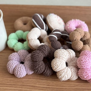 Op de afbeelding: Een verzameling handgemaakte, gebreide donuts in verschillende kleuren en stijlen. De donuts zijn onder meer lichtbruin, met chocolade glazuur, roze glazuur met hagelslag en een groene donut. De donuts zijn gerangschikt op een houten oppervlak.