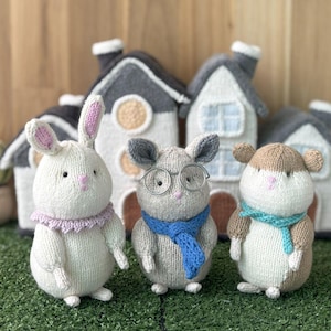 May include: Three knitted animal figures: a white bunny with pink ears and a purple collar, a gray mouse with glasses and a blue scarf, and a brown and white hamster with a teal scarf. Behind them are miniature houses.
