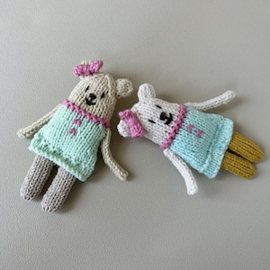 Peut inclure: Deux ours en peluche tricotés portant des robes bleu clair avec des bordures roses et des nœuds roses. Les ours ont des yeux bruns et des nez noirs.