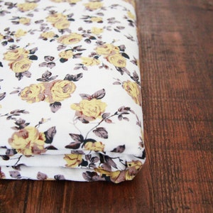 Peut inclure: Tissu plié avec un fond blanc et un motif floral répété de roses jaunes et de feuilles marron. Le tissu est plié sur lui-même, et le fond est une surface en bois brun foncé.