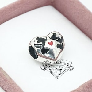 May include: A silver charm in the shape of a heart with black and red enamel detailing. The charm is in a white box with a pink interior.