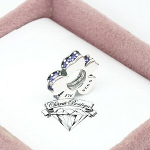 Puede incluir: Un charm de plata en forma de trébol de cuatro hojas con piedras de circonita cúbica de color púrpura. El charm está en una caja blanca con un interior rosa. La caja tiene el texto "Charm Boutique" en ella.