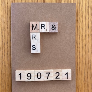 May include: A brown card with the letters "MR & MRS" spelled out with wooden letter tiles. The number "190721" is spelled out with wooden number tiles below the letters.