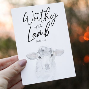 Könnte beinhalten: Eine weiße Karte mit dem Text "Worthy is the Lamb" in eleganter Schrift, darunter "Revelation 5:12". Am unteren Rand befindet sich eine Aquarellillustration eines Lammes. Die Karte wird vor einem verschwommenen, natürlichen Hintergrund gehalten.