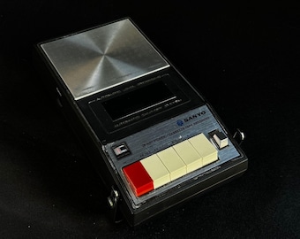 May include: A black Sanyo cassette tape recorder featuring a brushed silver metal top panel, a black cassette compartment, and a control panel with four off-white rectangular buttons and one red button, labeled 3-WAY POWER/CASSETTE TAPE RECORDER.