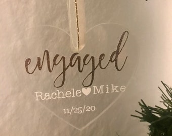 Może przedstawiać: Przezroczysta ozdoba w kształcie serca ze słowem "engaged" w kursywie. Poniżej imiona "Rachele" i "Mike" oddzielone symbolem serca, z datą "11/25/20" poniżej. Ozdoba zawieszona jest na białej wstążce.