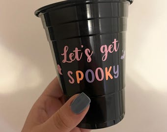 Pode incluir: Copo de plástico preto com as palavras "Let's get SPOOKY" em rosa, laranja e roxo. Um pequeno fantasma e um gráfico de morcego também estão incluídos. O copo é segurado por uma pessoa.