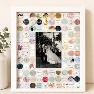 May include: A white rectangular picture frame containing a central black and white photograph of a couple in wedding attire, surrounded by a border of small, circular paper cutouts featuring various patterns, colors, and wedding-themed text.