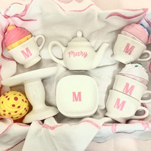 Puede incluir: Un juego de té de cerámica blanca con detalles en rosa, que incluye una tetera con la inscripción "Mary", tazas con la letra "M" y un pequeño soporte para pasteles. El juego está en una caja blanca con ribetes rosas.