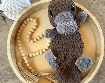 May include: A brown and gray crocheted plush toy shaped like a platypus. The toy is sitting in a wooden bowl with a string of wooden beads and tassels.