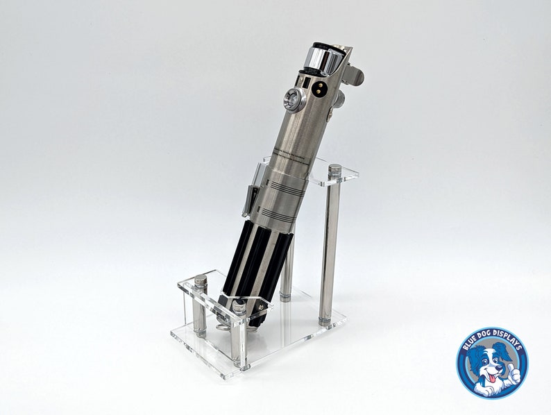 May include: A silver lightsaber replica displayed on a clear acrylic stand. The stand has two vertical supports and a base. The lightsaber has a black handle and a silver blade.