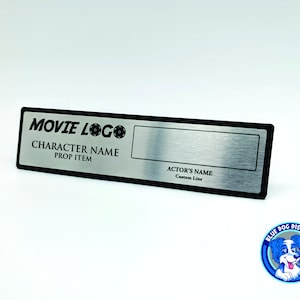 Może przedstawiać: Srebrna metalowa tabliczka z czarną ramką i tekstem "MOVIE LOGO", "CHARACTER NAME", "PROP ITEM", "ACTOR'S NAME" i "Custom Line".