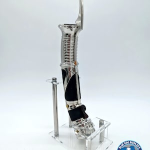 May include: A silver lightsaber prop with a black handle and a clear acrylic display stand. The stand has the text "Blue Dog Displays" on it.