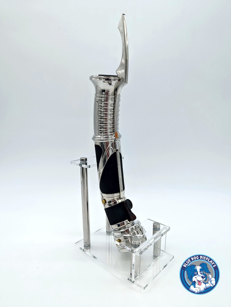 May include: A silver lightsaber prop with a black handle and a clear acrylic display stand. The stand has the text "Blue Dog Displays" on it.