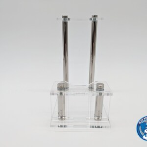 May include: A clear acrylic display stand with two silver metal rods. The stand is designed to hold two items, such as pens or pencils.