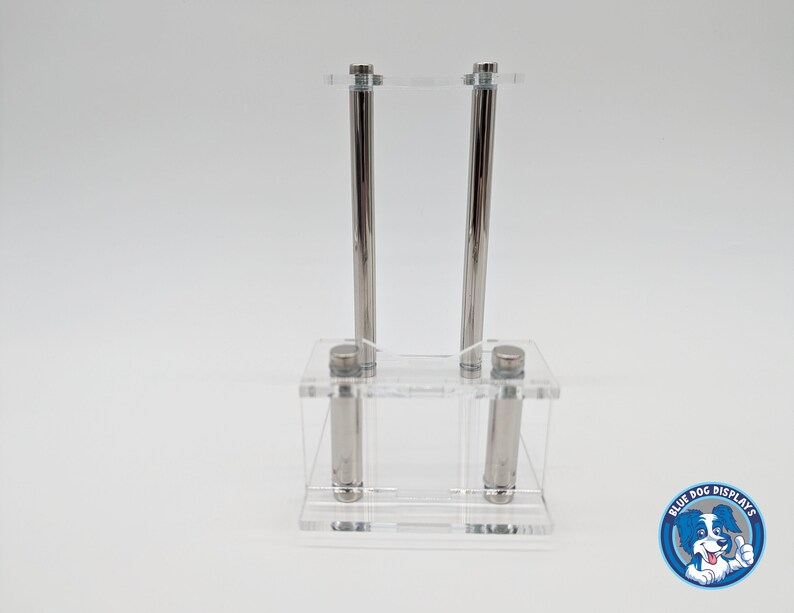 May include: A clear acrylic display stand with two silver metal rods. The stand is designed to hold two items, such as pens or pencils.