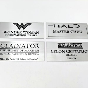 Op de afbeelding: Vier zilveren metalen plaquettes met zwarte tekst. De plaquettes tonen de namen van populaire fictieve personages en hun bijbehorende helmen. De namen zijn Wonder Woman, Master Chief, Gladiator en Cylon Centurion.