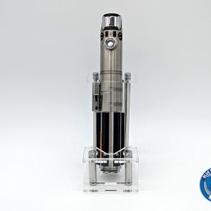 May include: A silver lightsaber replica displayed in a clear acrylic stand. The lightsaber has a black handle and a silver blade. The stand has a clear base and four clear acrylic rods.
