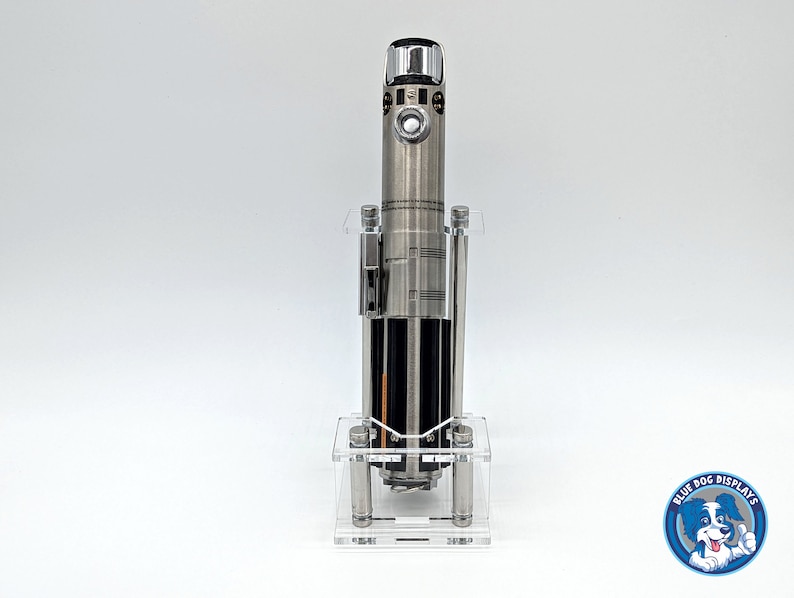 May include: A silver lightsaber replica displayed in a clear acrylic stand. The lightsaber has a black handle and a silver blade. The stand has a clear base and four clear acrylic rods.