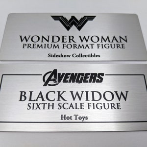 Puede incluir: Dos placas de metal plateado con texto negro. La placa superior presenta el logotipo de Wonder Woman y el texto "WONDER WOMAN PREMIUM FORMAT FIGURE Sideshow Collectibles". La placa inferior presenta el logotipo de los Vengadores y el texto "AVENGERS BLACK WIDOW SIXTH SCALE FIGURE Hot Toys".