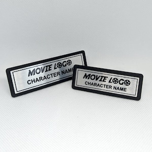 May include: Two rectangular name badges with the text "MOVIE LOGO" and "CHARACTER NAME" in black lettering. The badges have a silver-colored center and a black border. One badge is larger than the other.