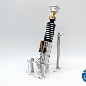 May include: A silver and gold lightsaber replica on a clear acrylic stand. The lightsaber has a black and silver handle with a gold top. The stand has two clear acrylic rods.