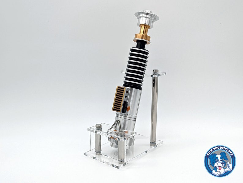May include: A silver and gold lightsaber replica on a clear acrylic stand. The lightsaber has a black and silver handle with a gold top. The stand has two clear acrylic rods.