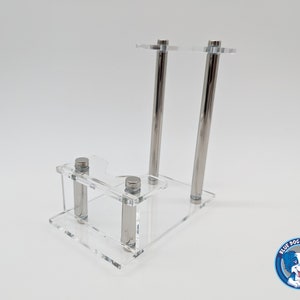 May include: A clear acrylic display stand with two silver metal rods. The stand has a small shelf and a larger top shelf. The stand is designed to hold a small object, such as a figurine or a collectible.