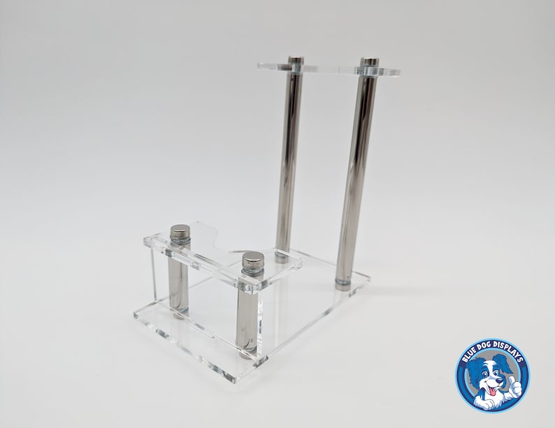 May include: A clear acrylic display stand with two silver metal rods. The stand has a small shelf and a larger top shelf. The stand is designed to hold a small object, such as a figurine or a collectible.