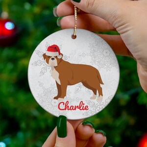May include: A round Christmas ornament featuring a brown and white dog wearing a Santa hat. The dog is standing on a white background with snowflake designs. The name "Charlie" is written in red script below the dog. The ornament has a gold string for hanging.
