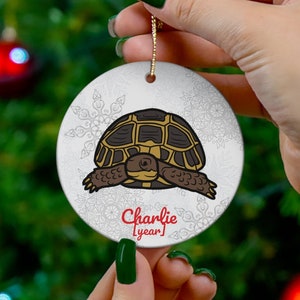 May include: A white ceramic Christmas ornament featuring a cartoon tortoise with a brown shell and a light brown body. The ornament has a gold string for hanging and the name "Charlie" is written in red below the tortoise.