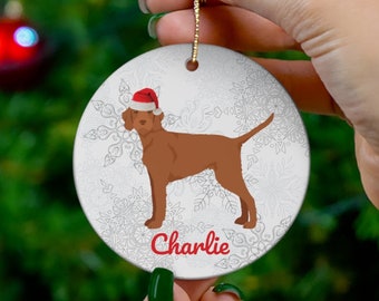 May include: A round, white Christmas ornament featuring a brown dog wearing a Santa hat. The dog is set against a backdrop of snowflakes. The name "Charlie" is written in red script below the dog. A gold string is attached for hanging.