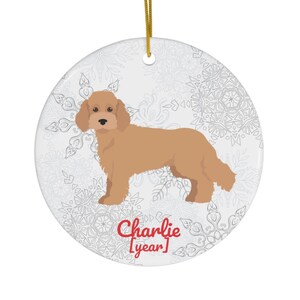 Op de afbeelding: Ronde keramische ornament met een illustratie van een bruine hond. Het heeft een gouden ophangkoord en een sneeuwvlokpatroon op een witte achtergrond. De tekst "Charlie [year]" is in het rood gedrukt.