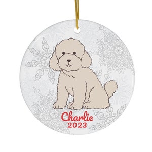 Può includere: Ornamento natalizio rotondo e bianco con un'illustrazione di barboncino marrone chiaro. L'ornamento ha un cordino dorato per appenderlo e un motivo a fiocco di neve. Il nome "Charlie" e l'anno "2023" sono stampati in rosso.