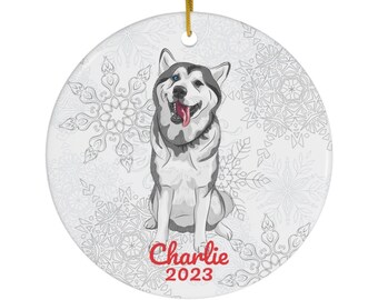 May include: A white ceramic Christmas ornament with a gray and white husky dog illustration. The dog is sitting with its tongue sticking out. The ornament has a white snowflake pattern and the text "Charlie 2023" in red.