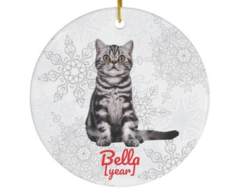 May include: Round ceramic ornament featuring a gray and black tabby cat with yellow eyes, sitting upright. The ornament has a gold string for hanging and a snowflake pattern background. The text "Bella [year]" is printed in red.