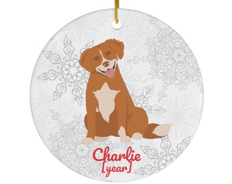 Könnte beinhalten: Runder Weihnachtsbaumschmuck aus Keramik mit einem Cartoon-Hund mit braunem und weißem Fell, der lächelt. Der Anhänger hat eine goldene Schnur zum Aufhängen und einen Hintergrund mit Schneeflockenmuster. Der Name "Charlie" ist rot geschrieben.