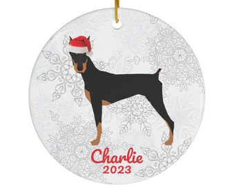 Op de afbeelding: Een witte keramische kerstversiering met een zwarte en bruine hond die een rode kerstmanmuts draagt. De versiering heeft een sneeuwvlokkenpatroon en de tekst "Charlie 2023" in rood.