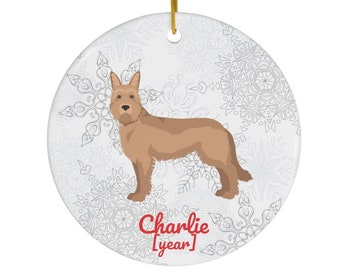 May include: A round, white ceramic ornament with a tan dog illustration. The ornament has a gold string for hanging and features a snowflake pattern. The text "Charlie [year]" is printed in red.