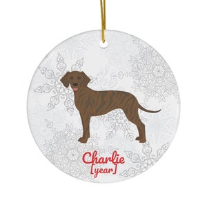 May include: Round ceramic ornament featuring a brown dog illustration with a striped pattern. The ornament has a gold string for hanging and a snowflake design on a white background. The text "Charlie [year]" is printed in red.