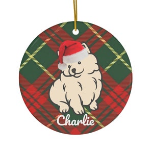 Pode incluir: Enfeite de Natal redondo com um cão creme usando um gorro de Papai Noel. O enfeite tem um fundo com padrão xadrez vermelho e verde. O nome do cão, "Charlie", está escrito em branco.