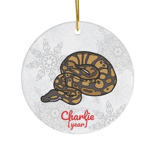 Op de afbeelding: Ronde, witte keramische ornament met een afbeelding van een bruine en beige slang. Het ornament heeft een gouden ophangkoord en de tekst "Charlie [year]" in het rood. Sneeuwvlokkenpatroon op de achtergrond.