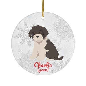 Puede incluir: Un perro blanco y marrón con un fondo de patrón de copo de nieve. El perro está sentado y el texto "Charlie [año]" está debajo del perro.