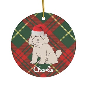 Può includere: Ornamento natalizio rotondo con un cane bianco con un cappello rosso di Babbo Natale. Il cane è seduto su uno sfondo a quadri verdi e rossi. Il nome "Charlie" è scritto in corsivo. Un filo d'oro è attaccato per appenderlo.