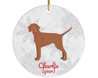May include: A round ceramic ornament with a white snowflake pattern. A brown dog with a red collar stands in the center of the ornament. The text "Charlie [year]" is printed in red at the bottom of the ornament.