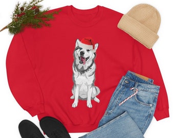 Puede incluir: Una sudadera roja con un gráfico de un husky con un gorro de Papá Noel. La imagen incluye un gorro beige, vaqueros azules, zapatos negros y una ramita de vegetación. Este es un artículo festivo de vacaciones.