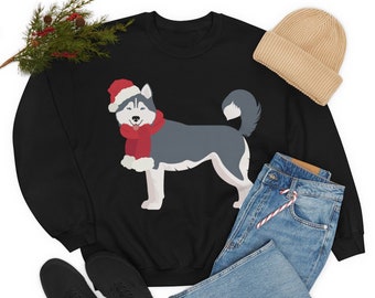 Puede incluir: Sudadera negra con un perro husky de dibujos animados con un gorro de Papá Noel y una bufanda roja. El perro es gris, blanco y negro. Un gorro beige, vaqueros azules y zapatos negros también están en la imagen. Una ramita de vegetación está en la esquina superior izquierda.