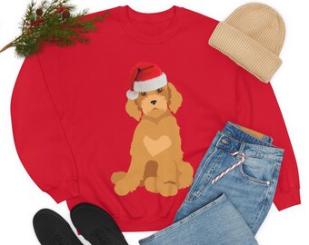 May include: A red sweatshirt featuring a cartoon dog wearing a Santa hat. The dog is light brown with a heart-shaped marking. The image also shows a beige beanie, blue jeans, black shoes, and a sprig of greenery.