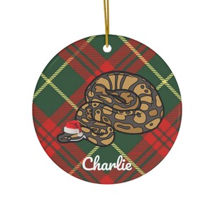 Peut inclure: Ornement de Noël rond en céramique avec un motif écossais vert, rouge et jaune. Un serpent de dessin animé portant un chapeau de Père Noël est enroulé sur le fond écossais. L'ornement porte le nom "Charlie" écrit en blanc en bas.